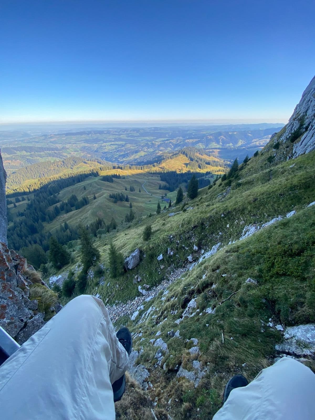 Blick über die eigenen Beine einen steilen Grashang hinunter ins weite Hügelland
