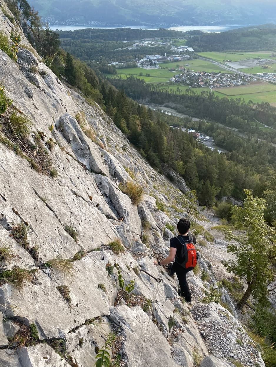 Kletterer mit rotem Rucksack in einer steilen Felswand hoch über dem Tal